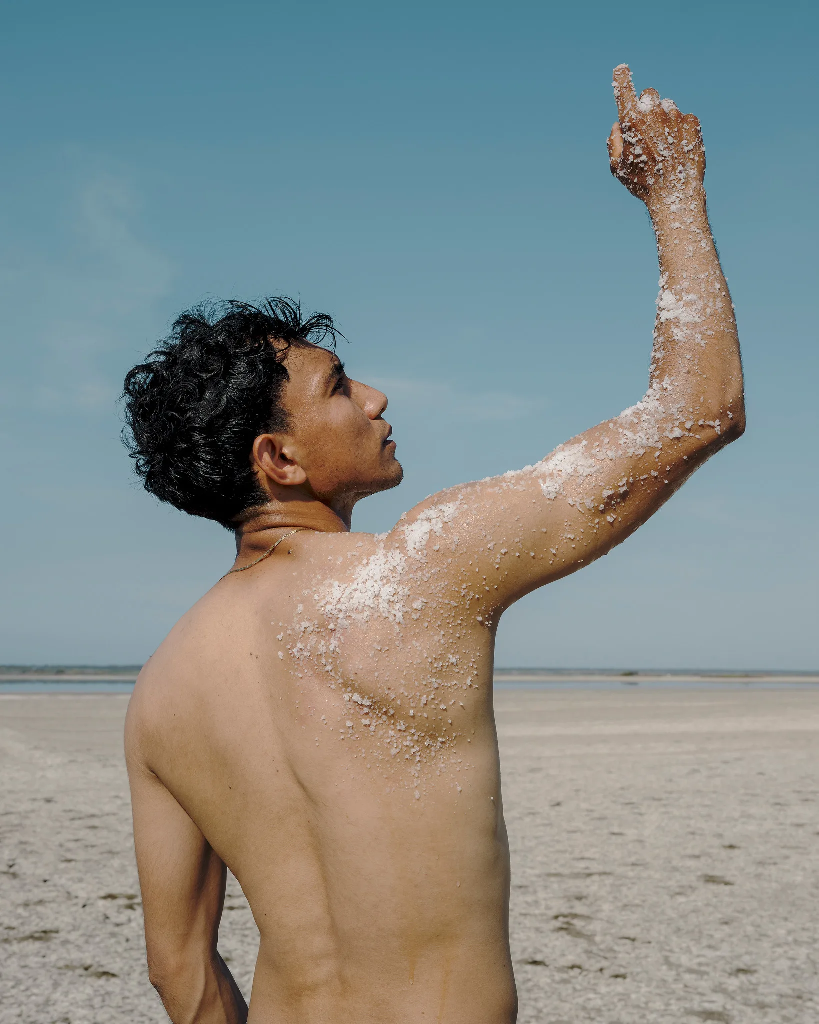 Joven de torso desnudo, de perfil y con la piel cubierta de costras de sal, alza el brazo derecho hacia el cielo en medio de un salar bajo un cielo azul despejado.