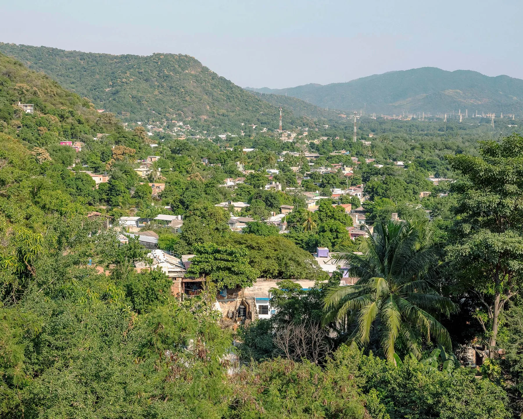 Vista elevada de un pueblo de casas bajas entre vegetación densa, con cerros arbolados al fondo y una palmera en primer plano.