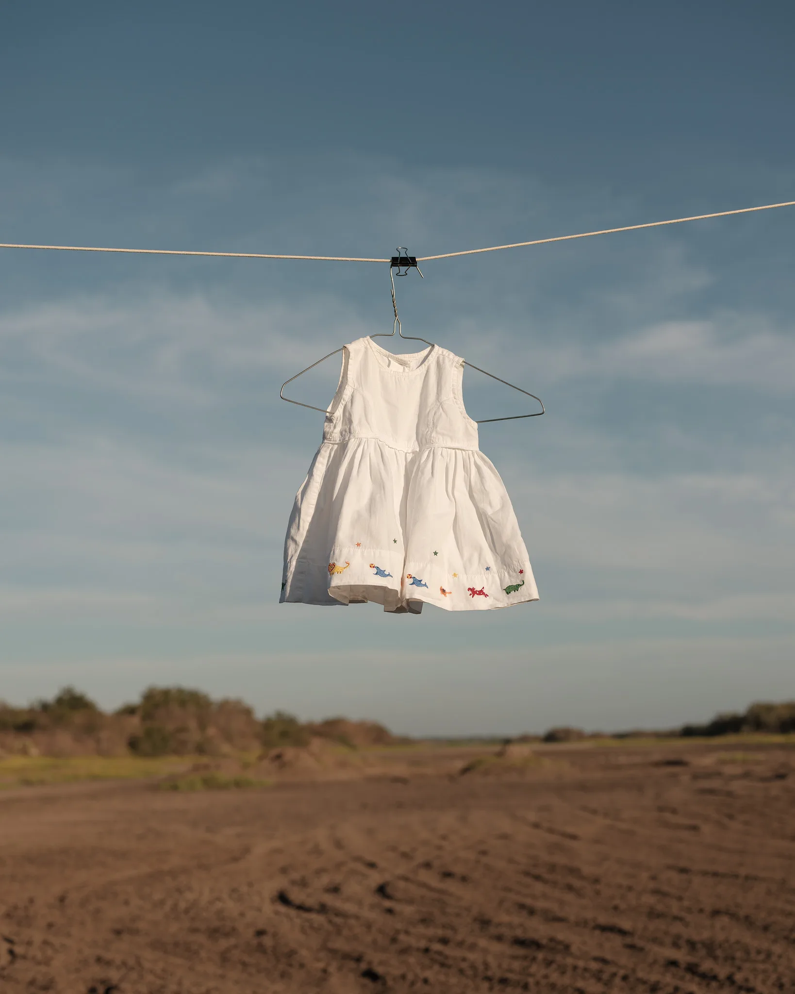 Vestido blanco de niña, bordado con animales de colores en el ruedo, colgado de un gancho en un tendedero sobre un terreno árido y cielo azul.