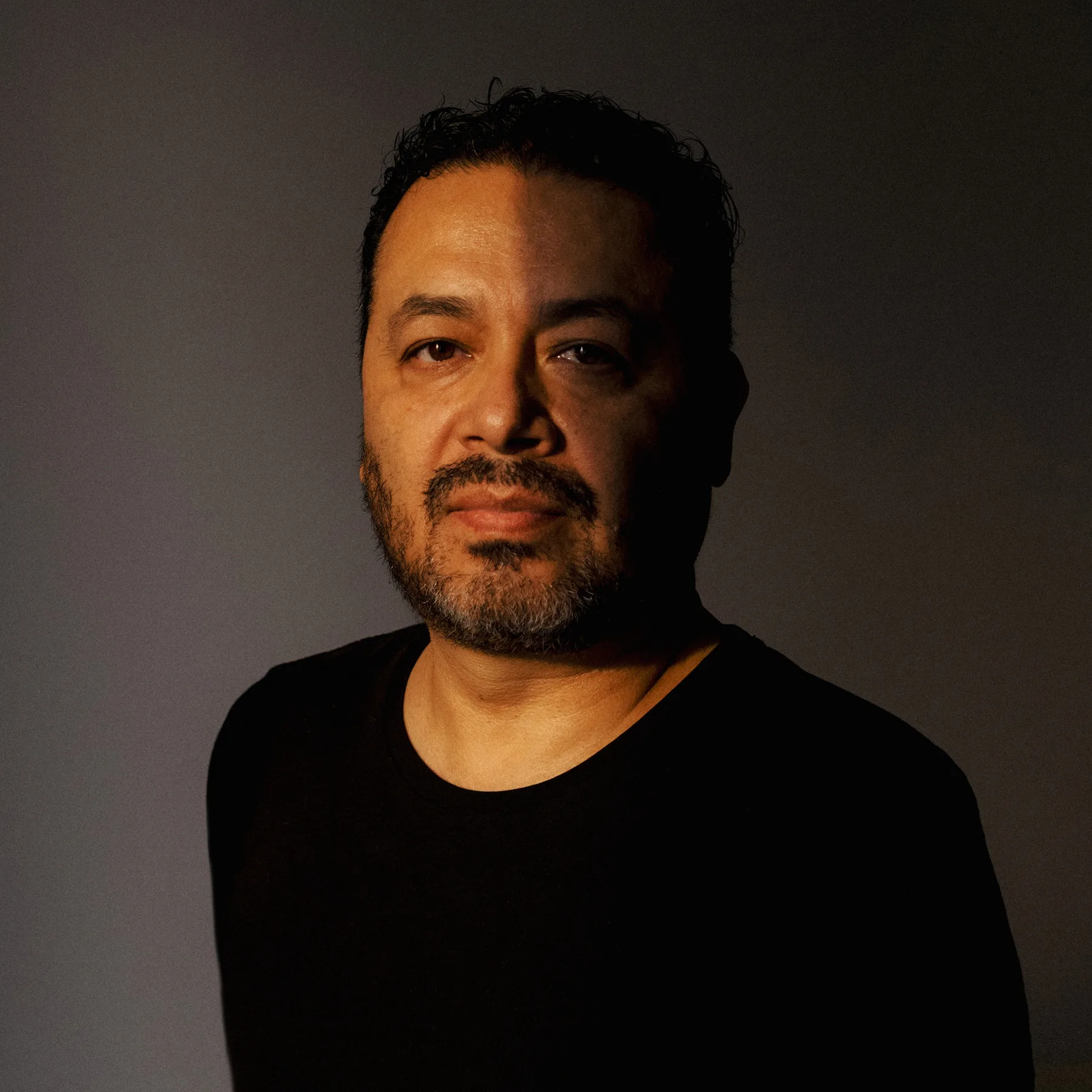 Retrato de Mario Alberto Chávez sobre fondo gris: hombre de barba entrecana y playera negra, iluminado de un costado con luz cálida, mirando a la cámara.