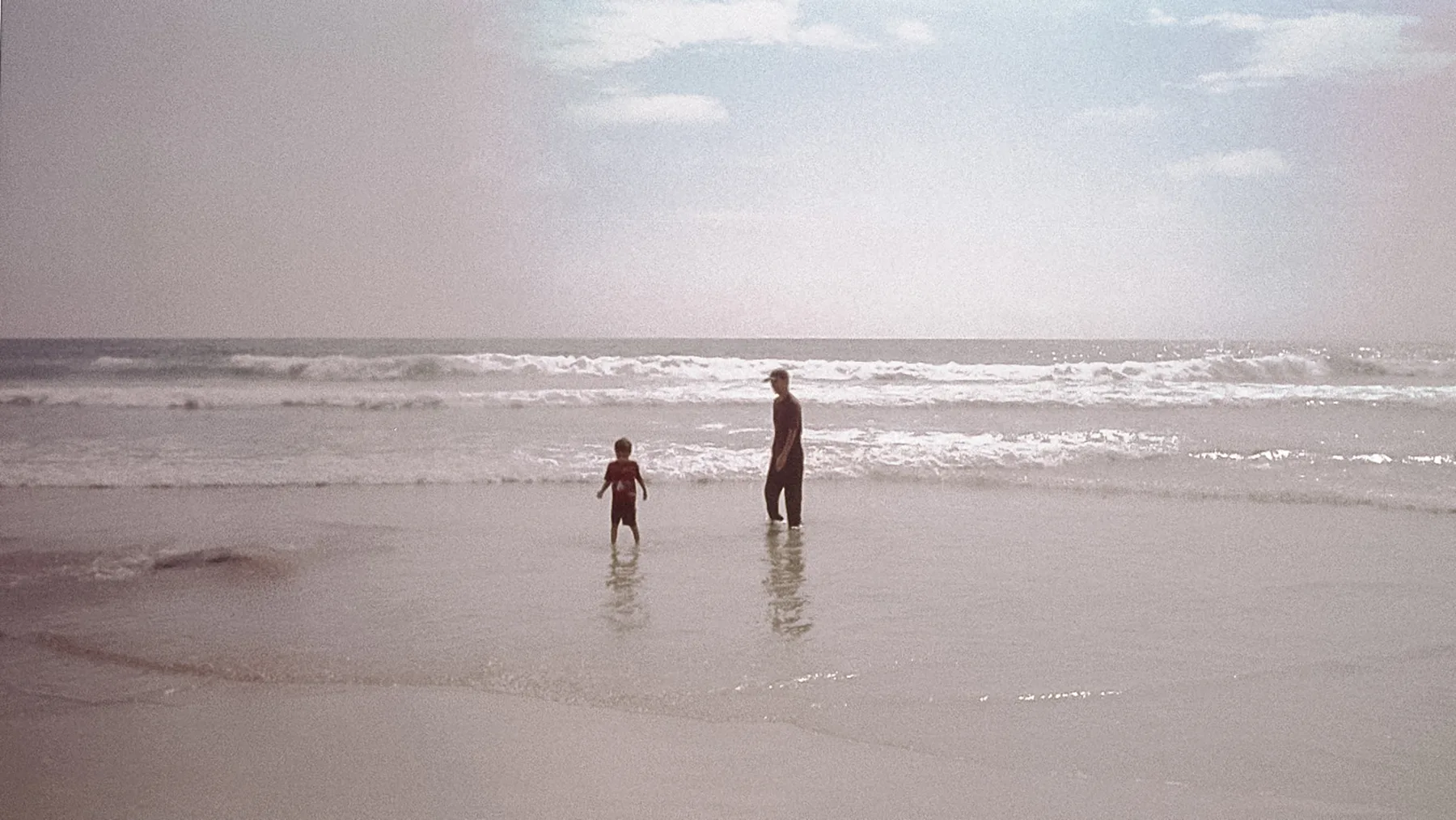 Fotografía analógica desvaída de un hombre y un niño de pie en la orilla del mar, de espaldas a la cámara, frente a las olas rompiendo.